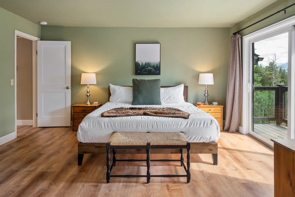 A serene and cozy king bedroom featuring a bed with a decorative throw, nightstands with lamps, and a large window that lets in natural light and offers a view of the outdoors. The décor exudes a mountain-chic vibe with premium bedding and thoughtful touches for relaxation.