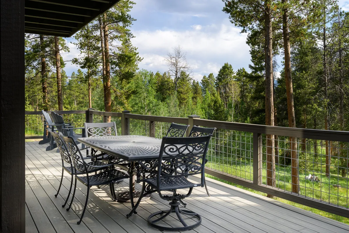 A peaceful exterior dining area on a wooden deck, surrounded by tall trees. An elegant table is set for outdoor meals, creating a perfect spot for gatherings or quiet moments in nature. The golden light of sunset illuminates the space, enhancing the sense of tranquility and exclusivity.