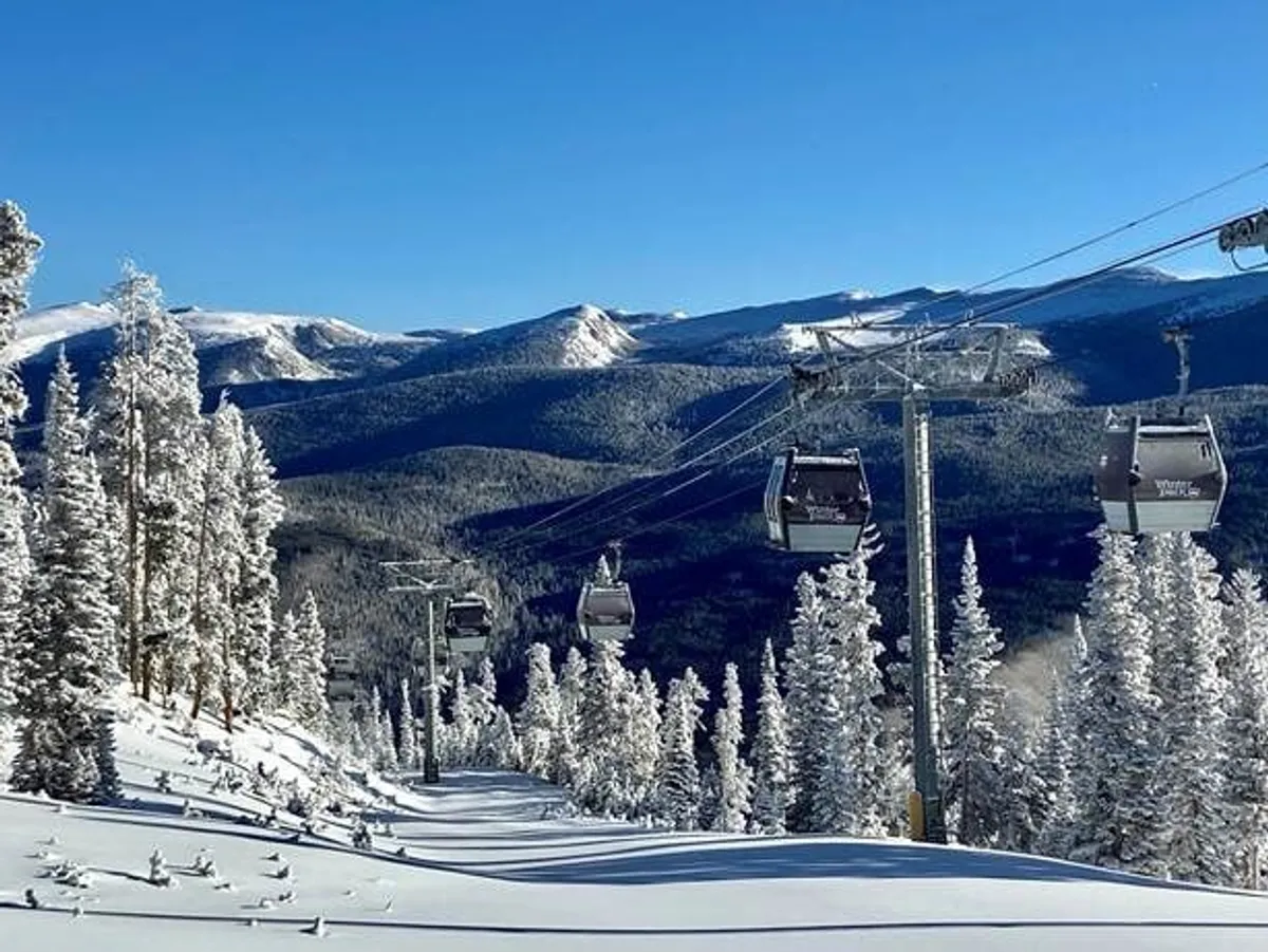 Ski lift gondolas glide above snow-covered trees, with a majestic mountain range as the backdrop. The snowy landscape is alive with the promise of winter sports, like skiing and snowboarding, and invites exploration of the local trails.