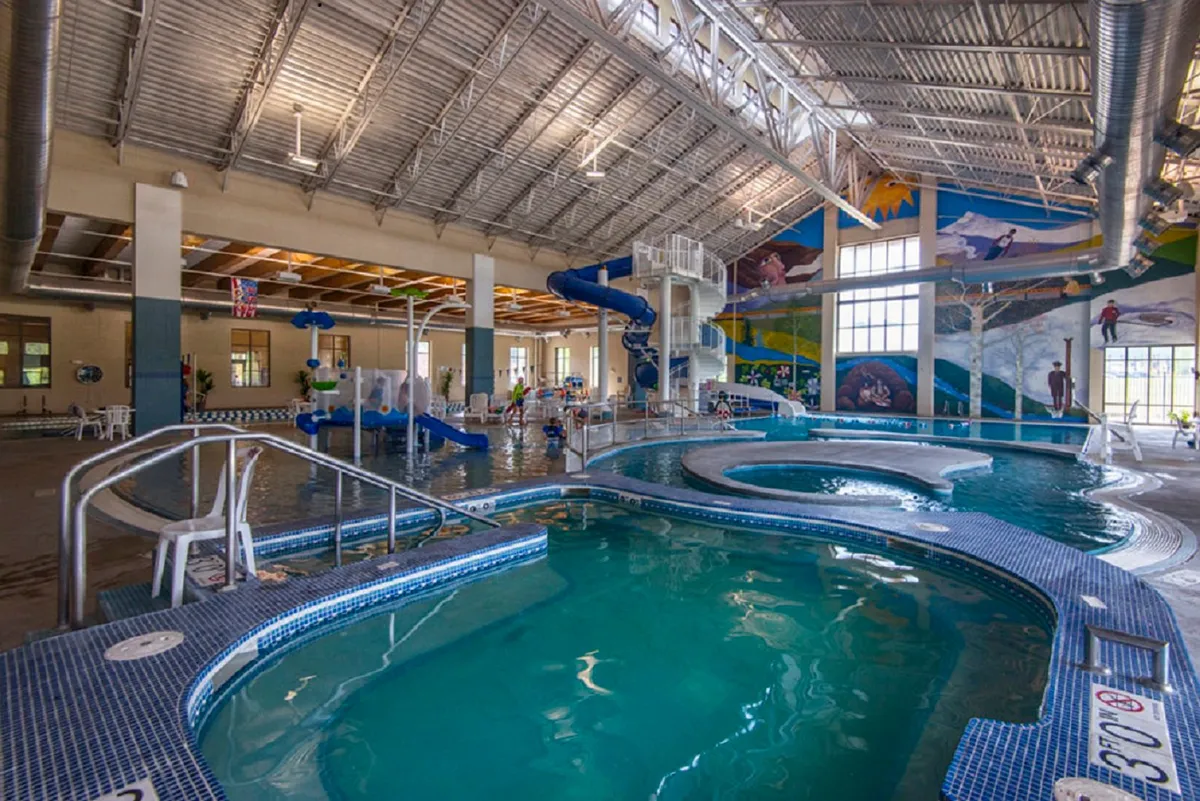 An interior view of a recreation center pool area, featuring colorful water slides, a spacious swimming pool, and vibrant murals. Children and families are depicted playing and enjoying aquatic activities, capturing the spirit of indoor fun mentioned in the section.