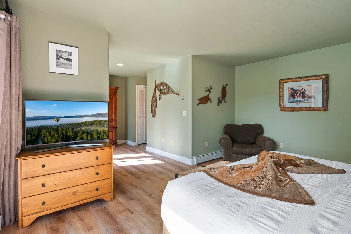 A warm, inviting bedroom with a bed featuring a decorative throw, a television displaying a nature scene, a wooden dresser, artwork, and an armchair. The room is designed for relaxation, with both natural light and entertainment options.
