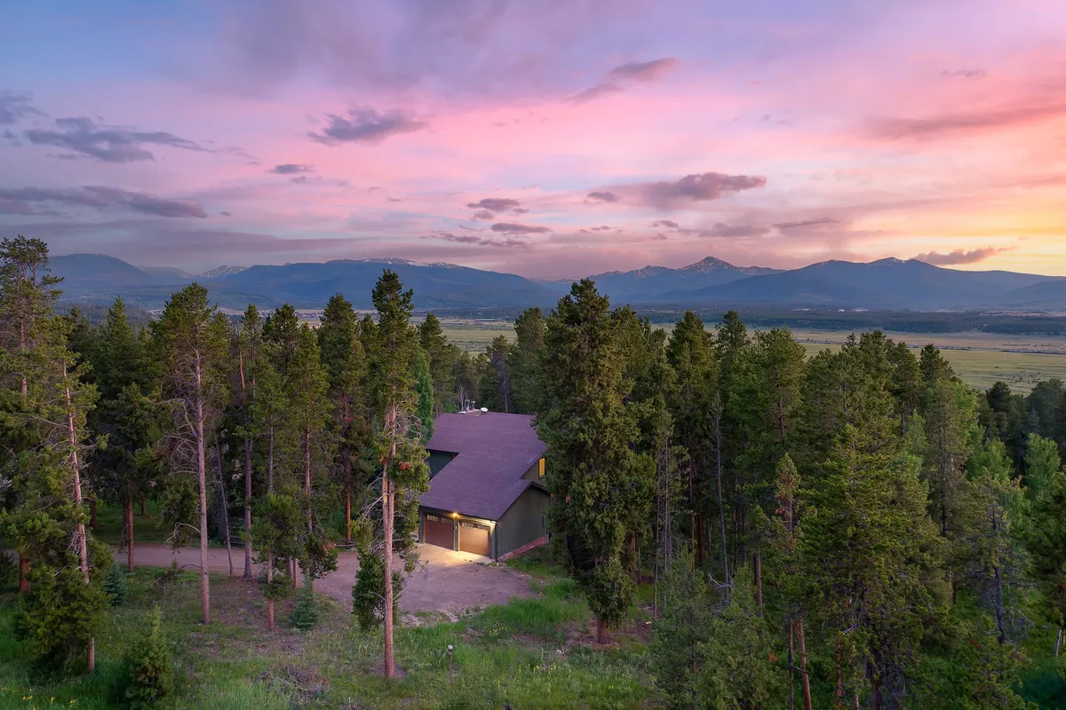 The house set against a stunning mountain backdrop with colorful skies, surrounded by tall trees, and a spacious yard visible. The exterior exudes tranquility, with cues to the natural adventure and exploration that awaits just beyond the home.