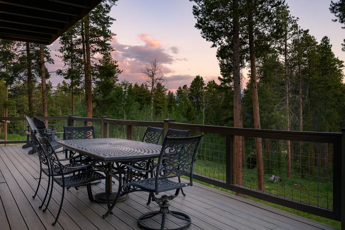 This image displays an inviting exterior patio with a table and metal chairs set against a tree line, under a colorful sunset sky. The setting is peaceful and perfect for outdoor relaxation or dining, with a sense of privacy and enclosure from the surrounding nature.
