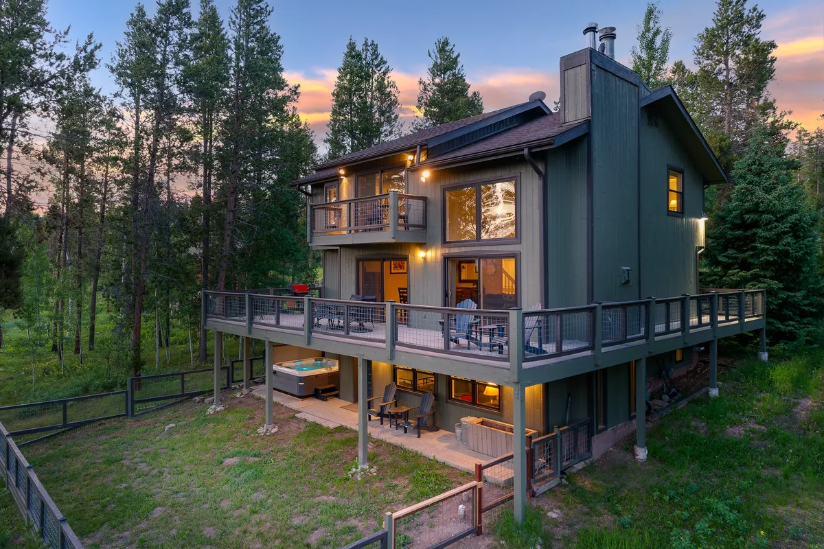The image showcases a beautiful two-story mountain house nestled among tall pines, featuring a spacious deck and an inviting hot tub. The deck is set up for outdoor relaxation and entertaining, hinting at sunset views and star-filled nights. The tranquil natural surroundings create a cozy, peaceful atmosphere ideal for gatherings and unwinding.
