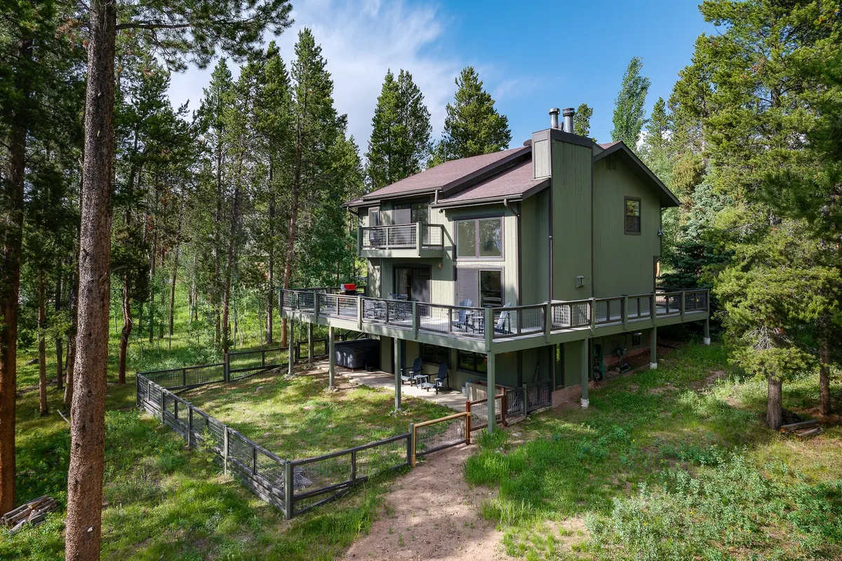The green exterior of the house sits harmoniously among towering trees, with a spacious deck extending out for gathering, relaxing, or stargazing. The setting is serene and inviting, ideal for returning after a day of adventure to unwind and connect with nature.