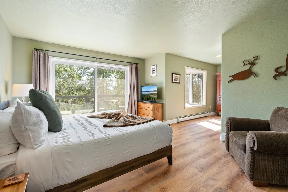 A tranquil king bedroom with a bed, windows streaming in sunlight, an armchair, wooden furniture, and decorative wall art. The space is inviting and comfortable, ideal for reading, relaxing, or enjoying peaceful mornings.