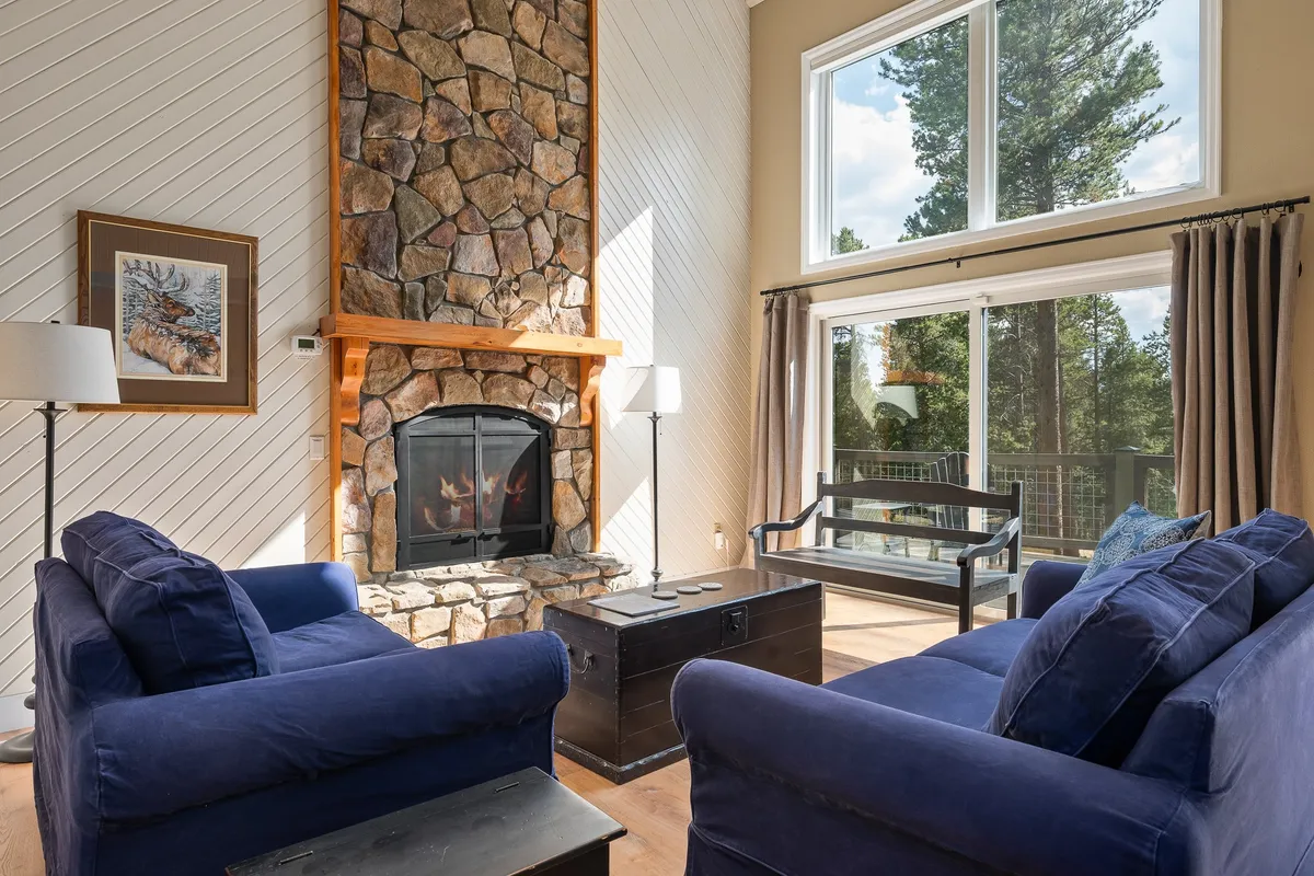 A spacious living room featuring a grand stone fireplace flanked by large panoramic windows that fill the space with natural light. Plush, comfortable seating is arranged around the fireplace, inviting guests to relax and enjoy the mountain views. The overall ambiance is warm, welcoming, and ideal for gathering with family or friends.