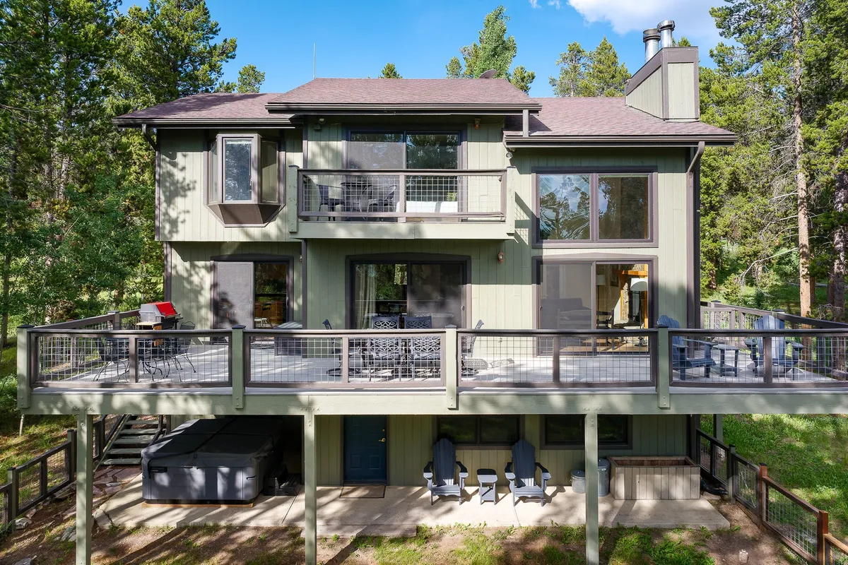 A spacious deck with a hot tub is surrounded by tall trees and features large windows that connect the interior with the outdoors. The scene invites relaxation and social gatherings, with ample room for a group to lounge or enjoy a barbecue together.