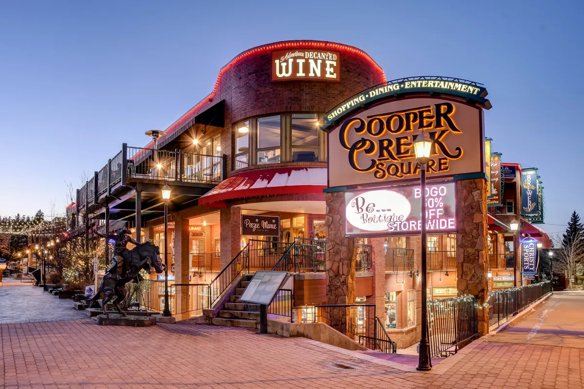 The Cooper Creek Square sign is visible, flanked by a restaurant and wine shop adorned with decorative lighting, signaling a lively atmosphere ideal for evening outings. The scene is festive, suggesting opportunities for dining, shopping, and entertainment just minutes from the lodge.